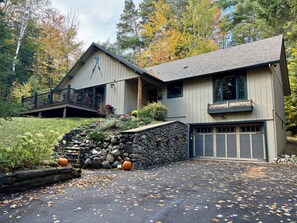 A two-story house with a stone retaining wall, a garage, and a deck with a railing. - Camp Kahuna - Near Mirror Lake - Dog Friendly (Lake Placid)
