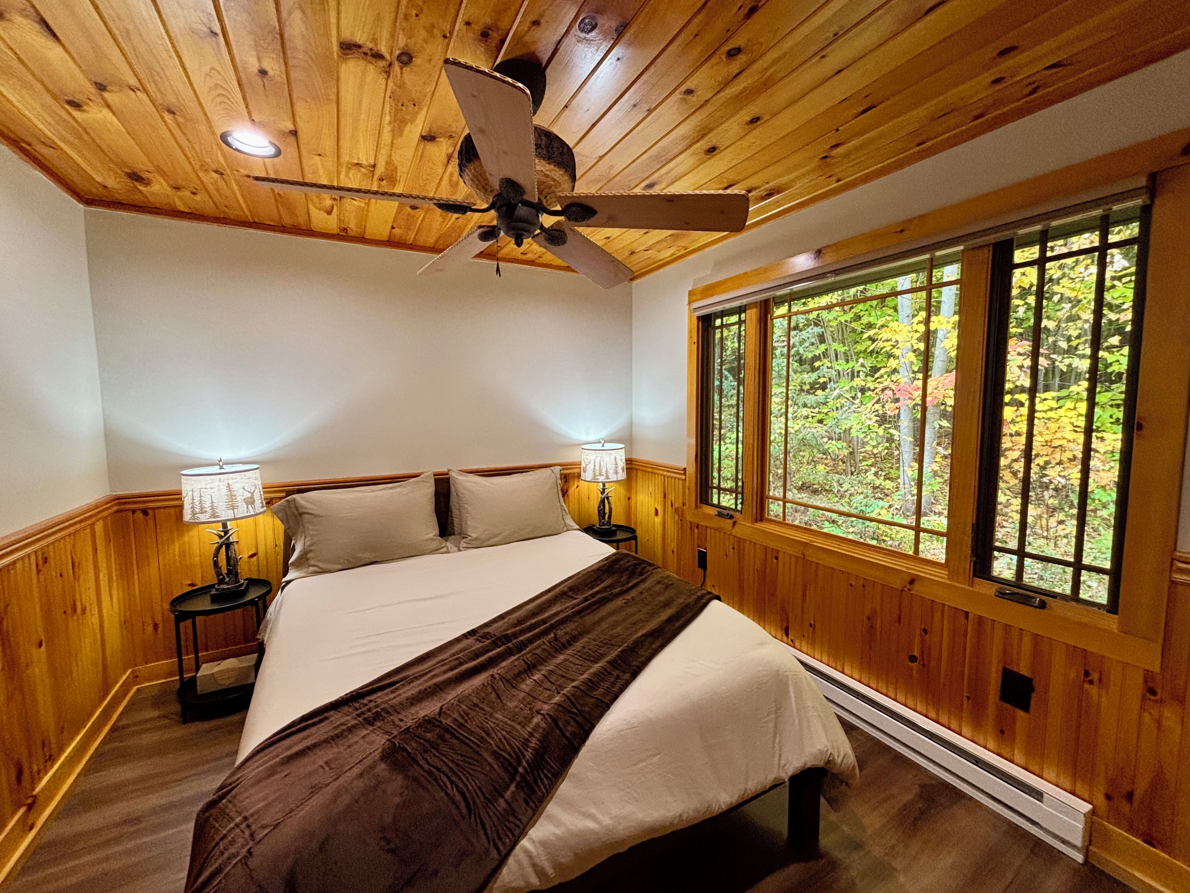 A bedroom with a wooden ceiling, a bed with a brown blanket, two bedside lamps, and a window with a view of trees.