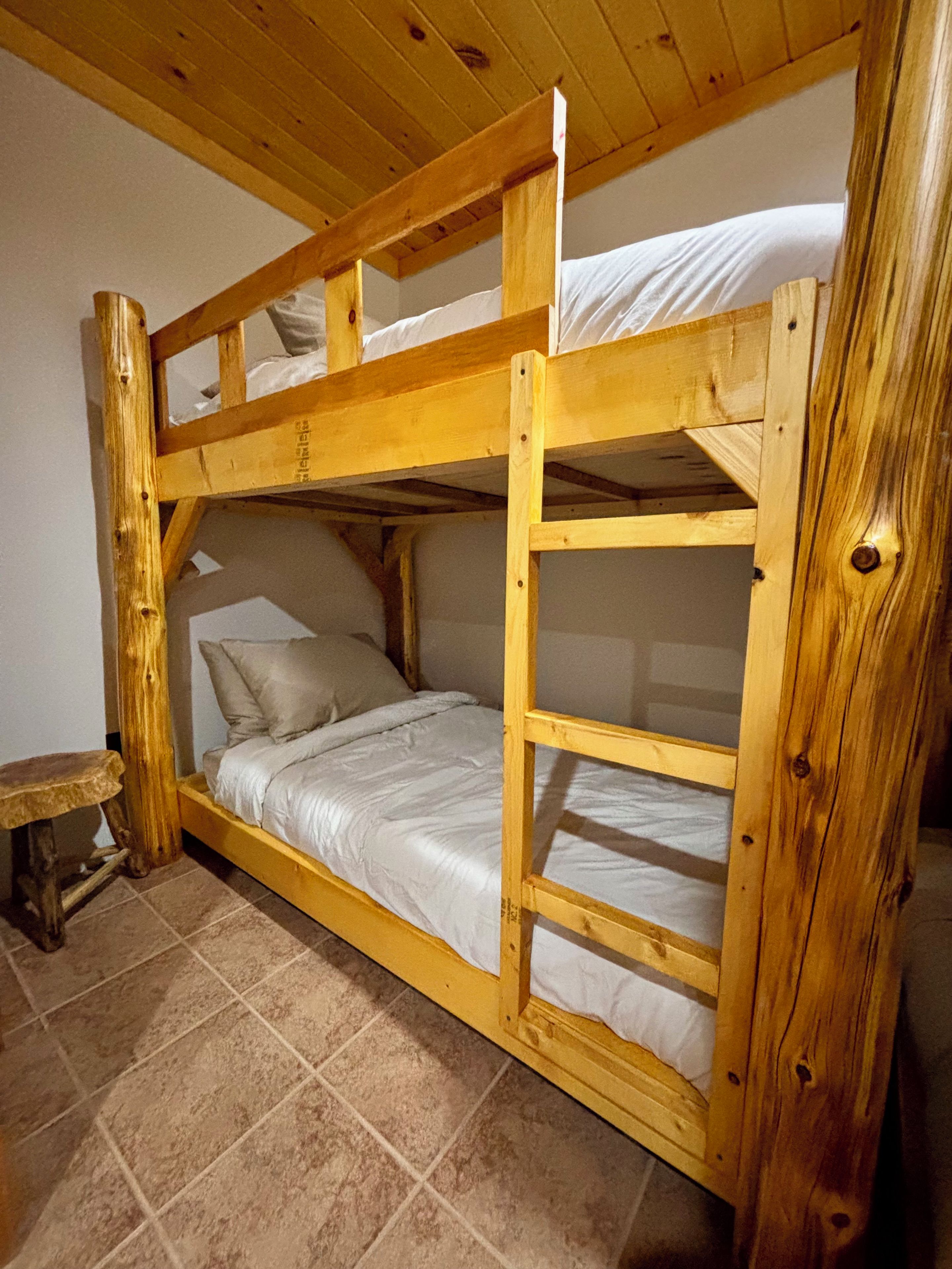 Een stapelbed met houten frame, een klein krukje en een betegelde vloer.