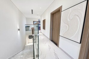 Een moderne hal met een glazen balustrade, een houten deur en een groot, gestructureerd kunstwerk aan de muur.