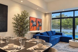 Uma área de refeições moderna com mesa posta para quatro pessoas, um sofá azul e uma janela grande com vista para uma piscina.