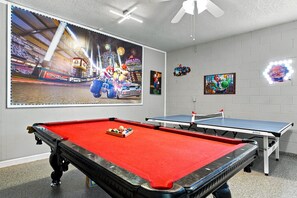 A pool table with a red felt surface and a set of pool balls, a second table with a blue felt surface, and a large poster of a racing game on the wall. - 6BR Windsor Hills Home Near Disney with Pool (Kissimmee)