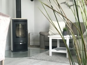 A modern living room with a black wood-burning stove, a grey sofa, a white coffee table, and a potted plant. - Little Baked Apple (Wrixum)