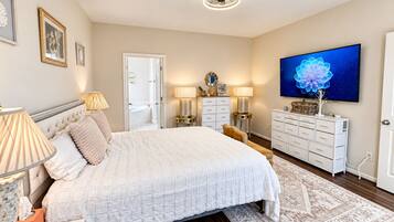A bedroom with a large bed, bedside lamps, a dresser, a television, and a bathroom visible through an open door.