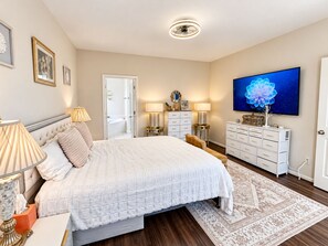 A bedroom with a large bed, bedside lamps, a dresser, a television, and a bathroom visible through an open door. - Elegant Private Master Bedroom Suite • Private Spa Bath, Soaking Tub & Patio (Houston)