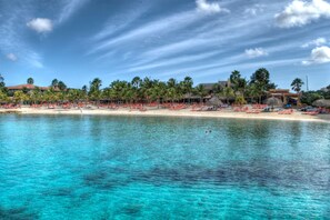 A beach resort with a clear blue sea, rows of red lounge chairs, palm trees, and a building with a red roof. - Poco Poco Villa at Blue Bay Resort | Pool, Beach & Golf (Willemstad)