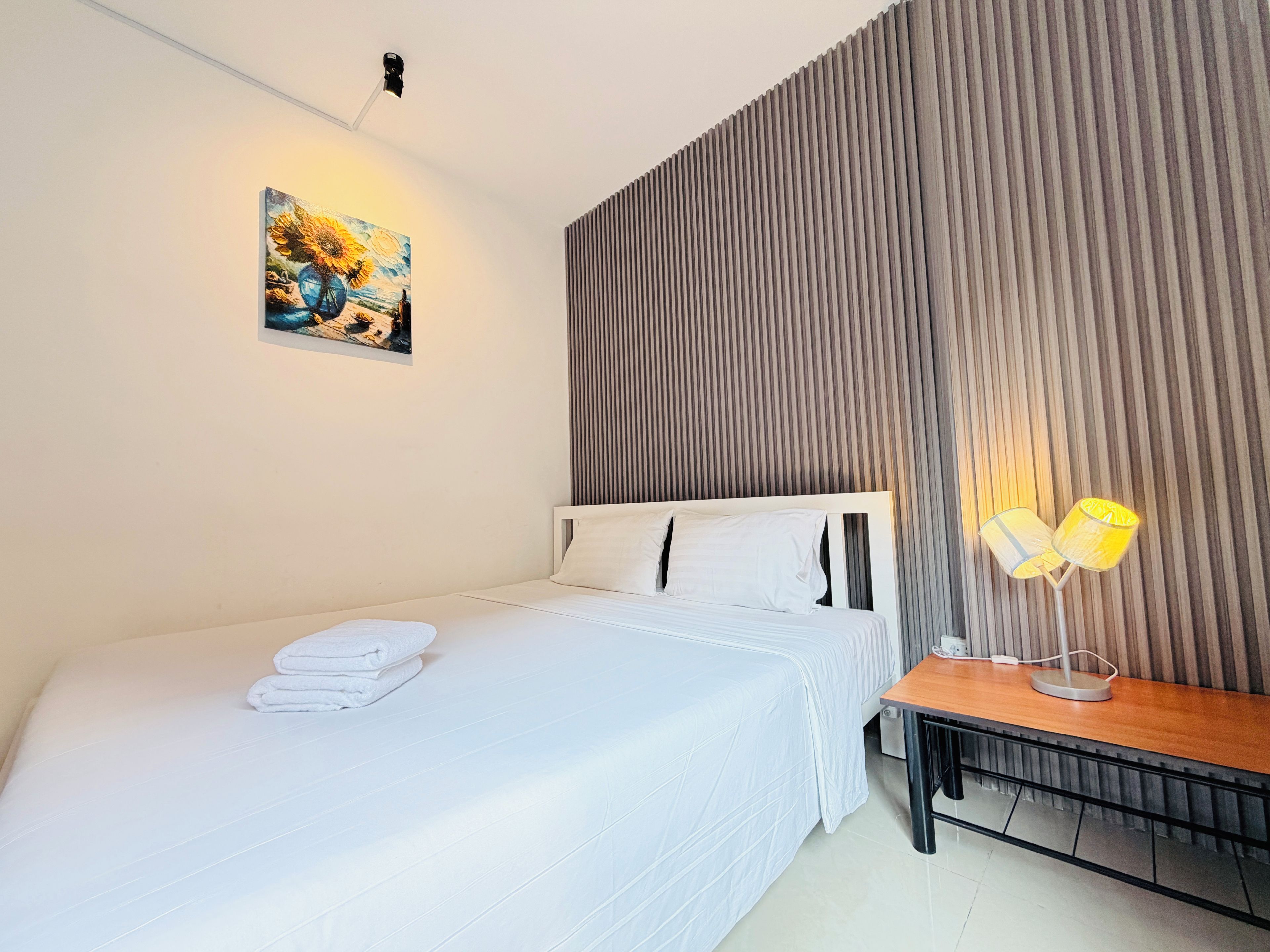A hotel room with a neatly made bed, a bedside table with a lamp, a wall-mounted artwork, and a textured wall panel.
