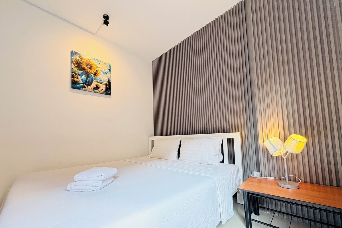 A hotel room with a neatly made bed, a bedside table with a lamp, a wall-mounted artwork, and a textured wall panel.