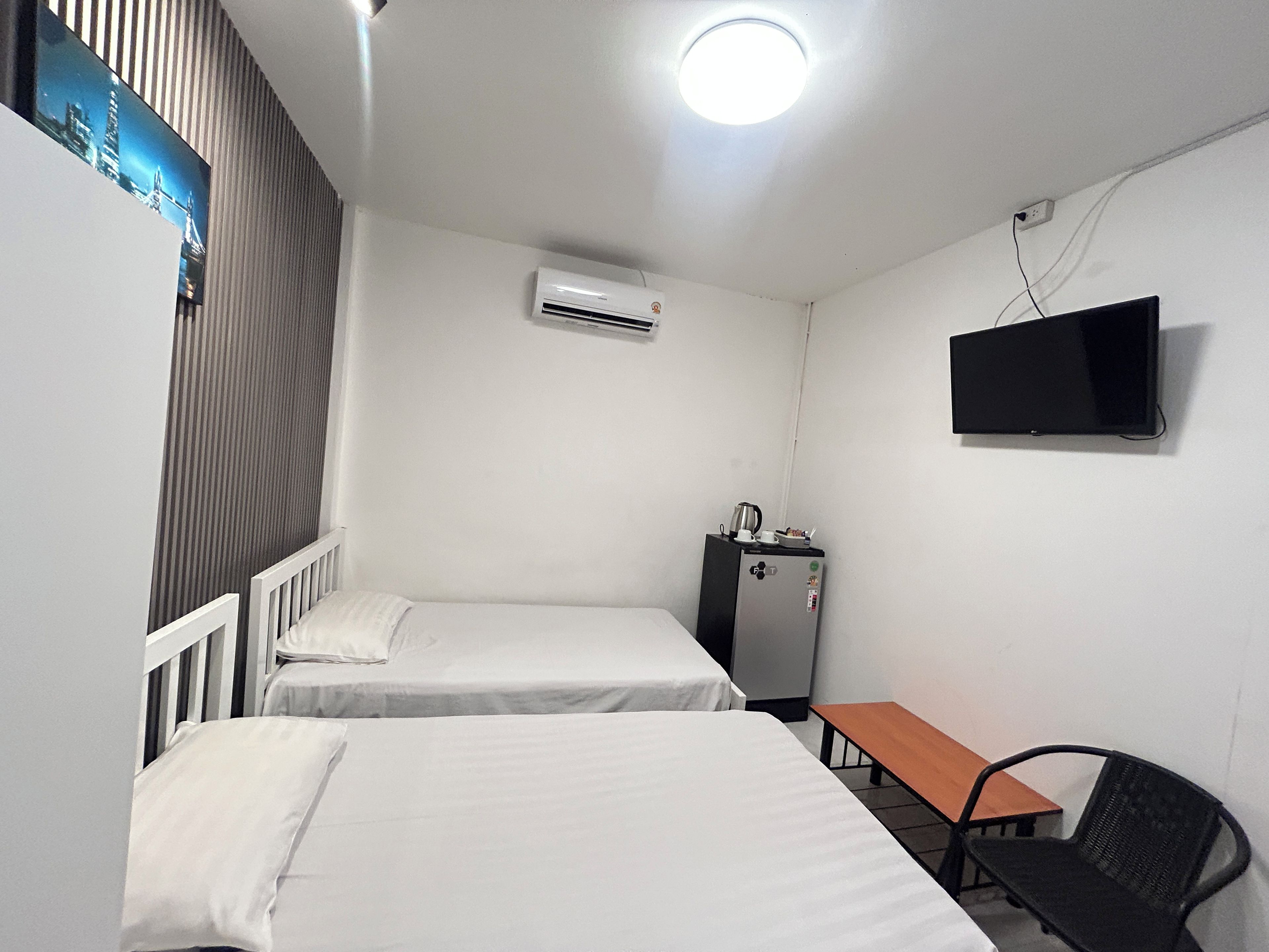 Ein Zimmer mit zwei Einzelbetten, einem kleinen Tisch, einem Sessel, einem an der Wand montierten Fernseher und einer Klimaanlage.