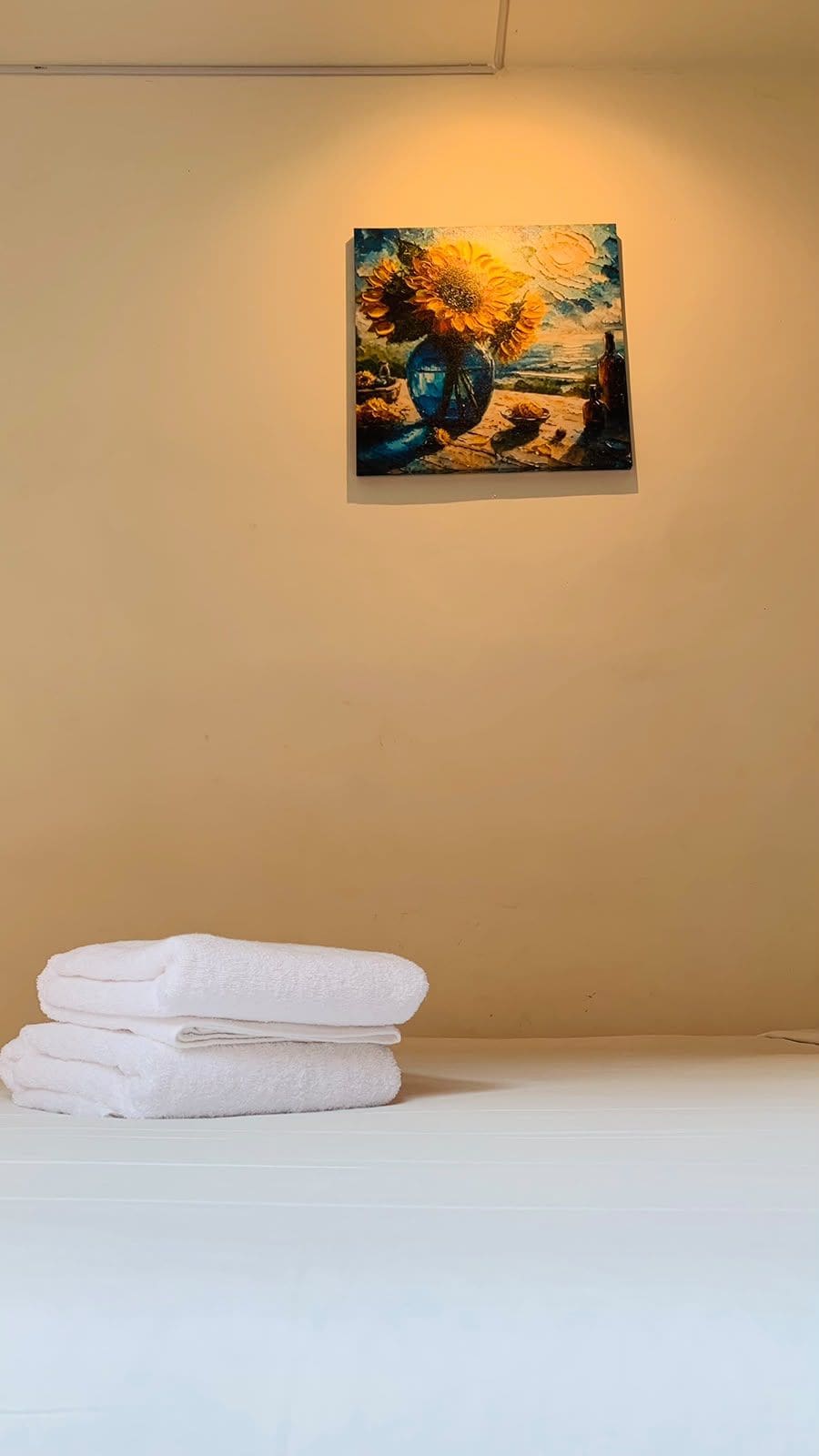 A painting of sunflowers and a blue vase on a wall, with a stack of white towels on a white surface.