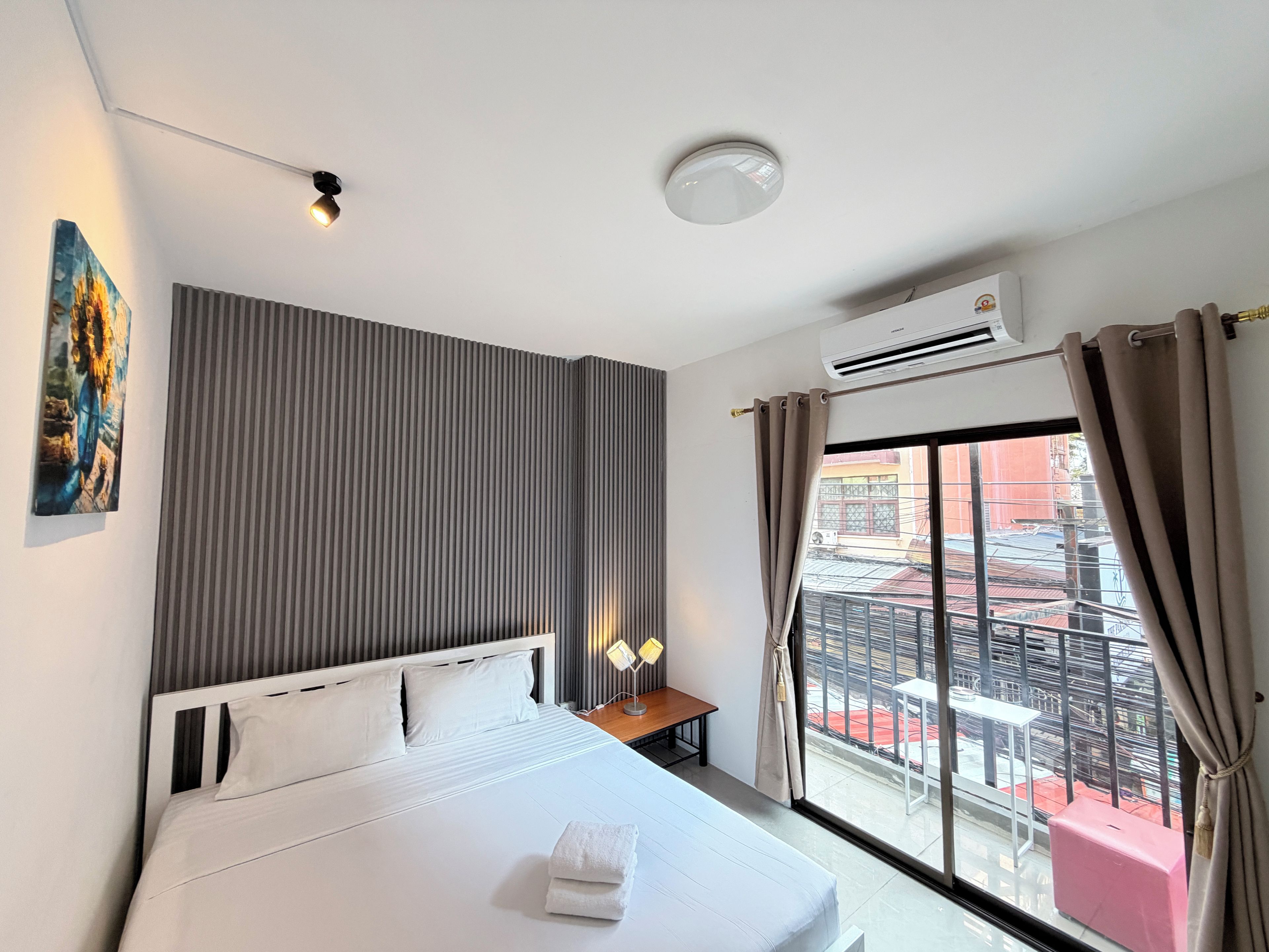 A hotel room with a bed, a bedside table with a lamp, a painting on the wall, and a view of the outside balcony.