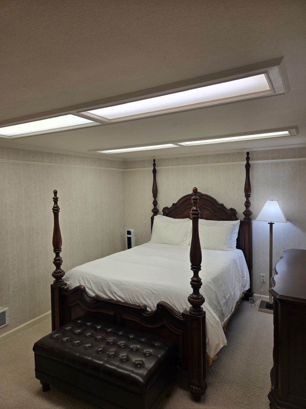 Una cama con dosel, un reposapiés capitonado, una lámpara de noche y un candelabro en el techo.
