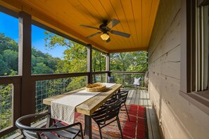 Una terraza de madera con mesa y sillas, un ventilador de techo y vistas al bosque.