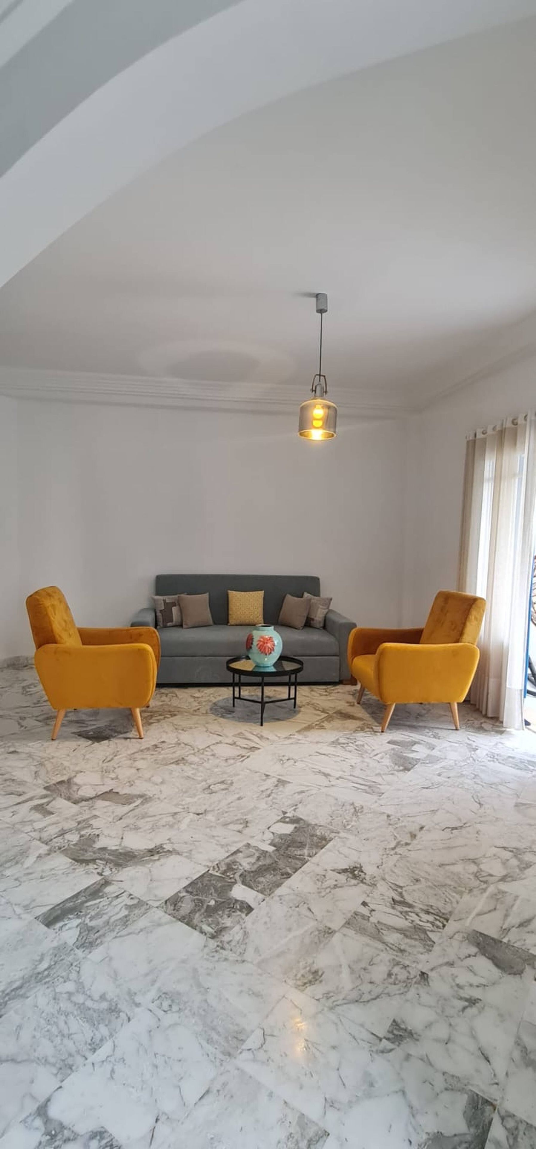 A living room with a marble floor, a grey sofa, two yellow armchairs, and a hanging light fixture.