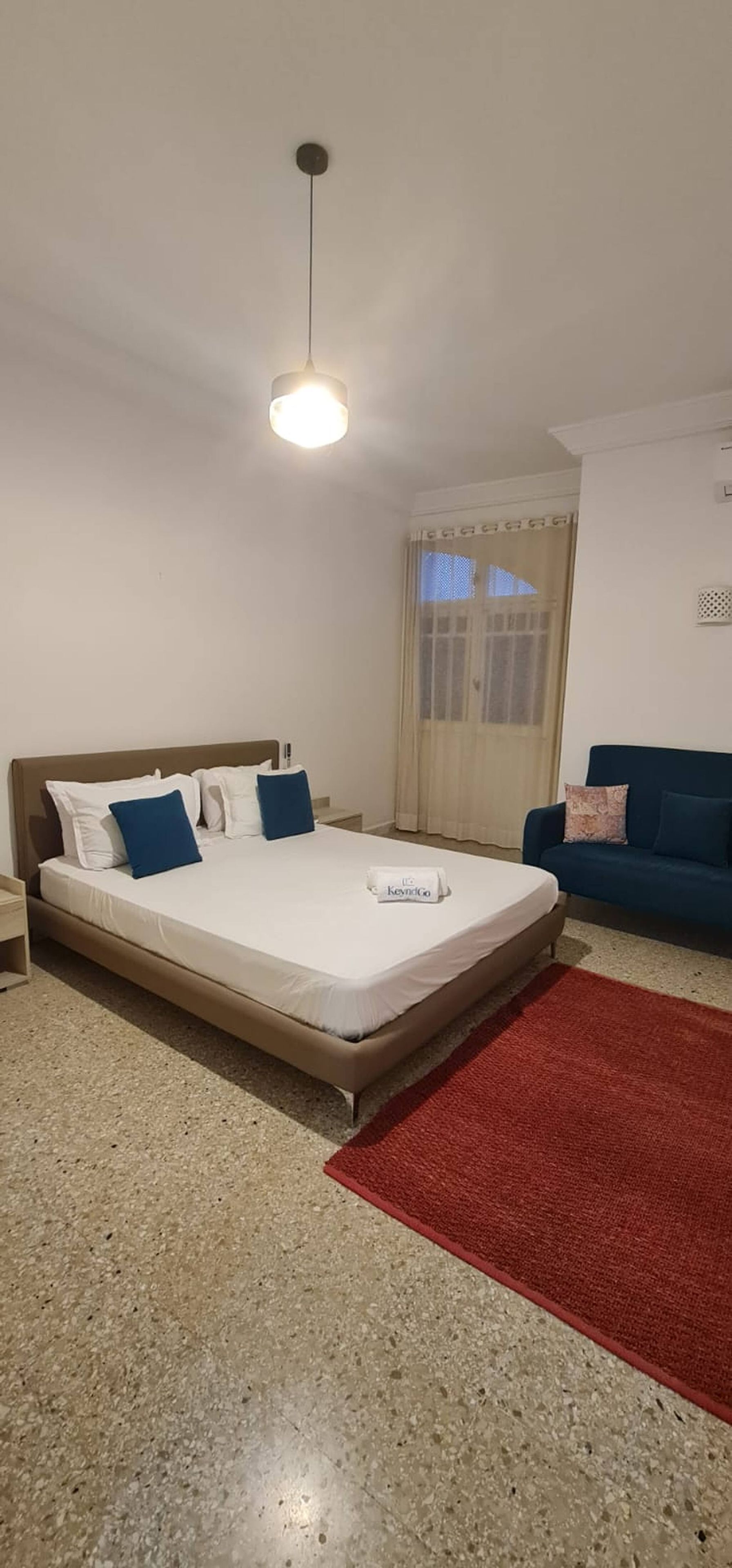 Una habitación de hotel con una cama, un tapete rojo, un sofá azul y una ventana con cortinas.