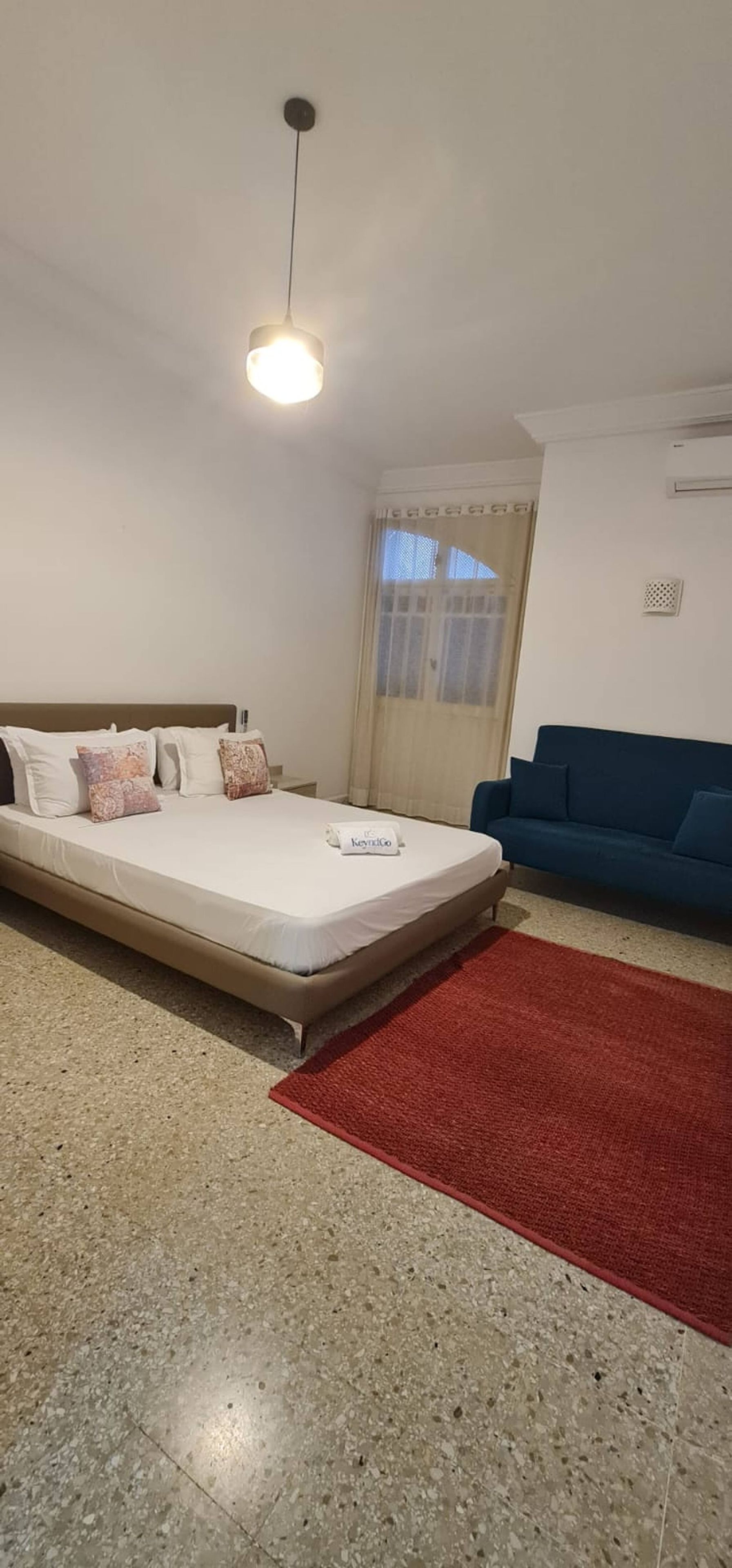 Un dormitorio con cama, un sofá azul, un tapete rojo y una ventana con cortinas.