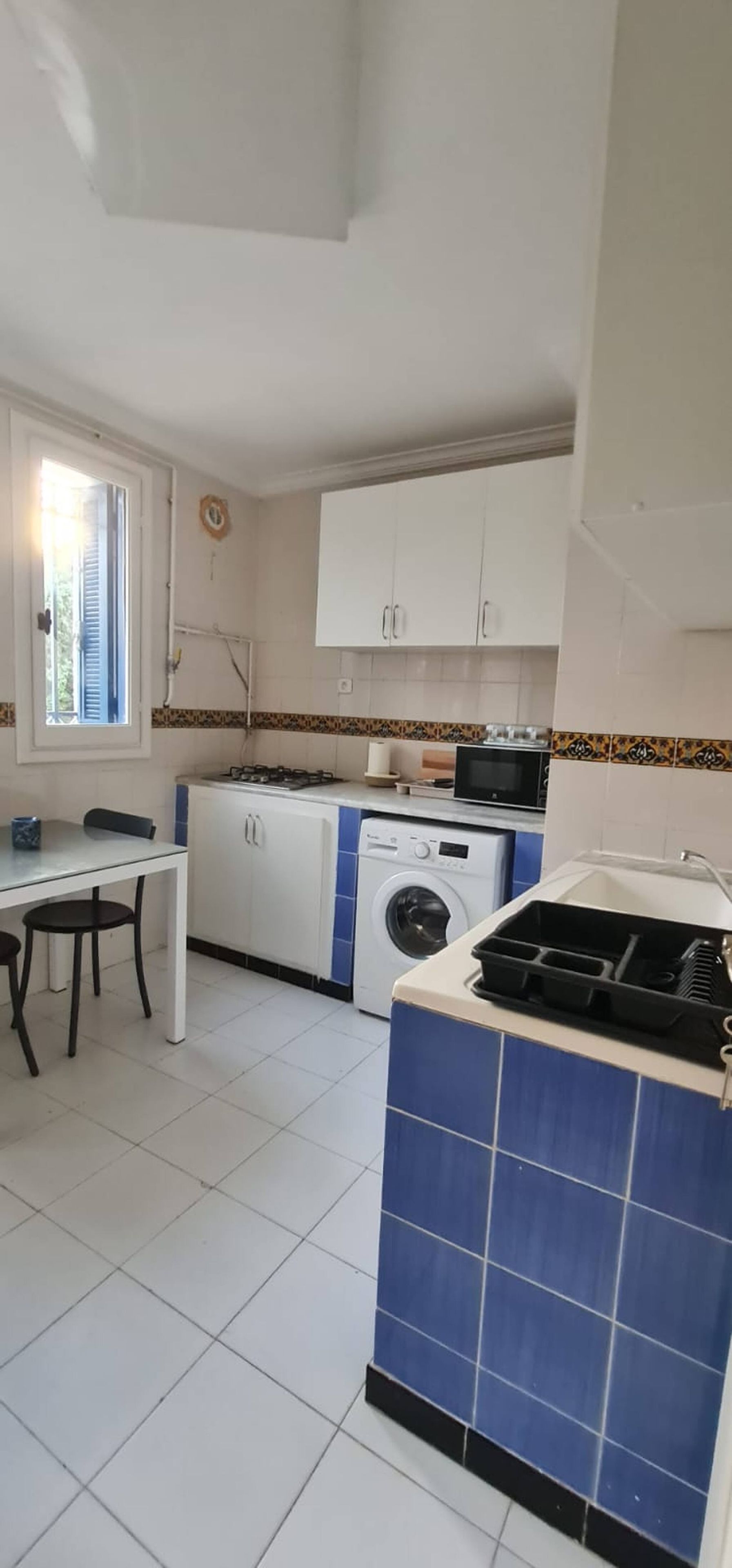 A compact kitchen with white cabinets, a washing machine, a microwave, and a dining area with a table and chairs.