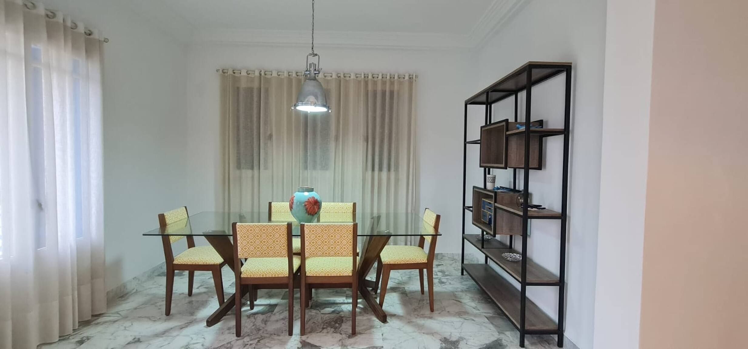 Un comedor con mesa de vidrio, cuatro sillas, un jarrón, una lámpara colgante y una estantería.