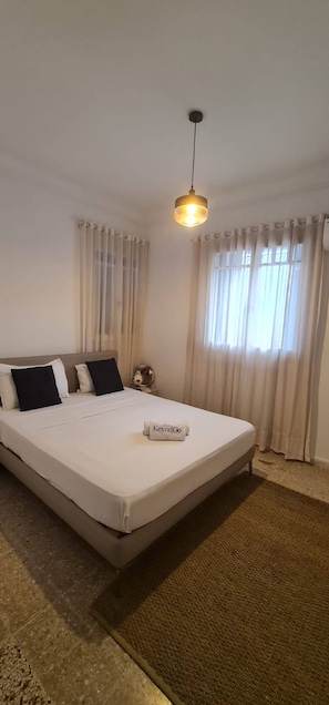 Una habitación de hotel con una cama, una lámpara colgante, una mesita de noche y un ventanal con cortinas.