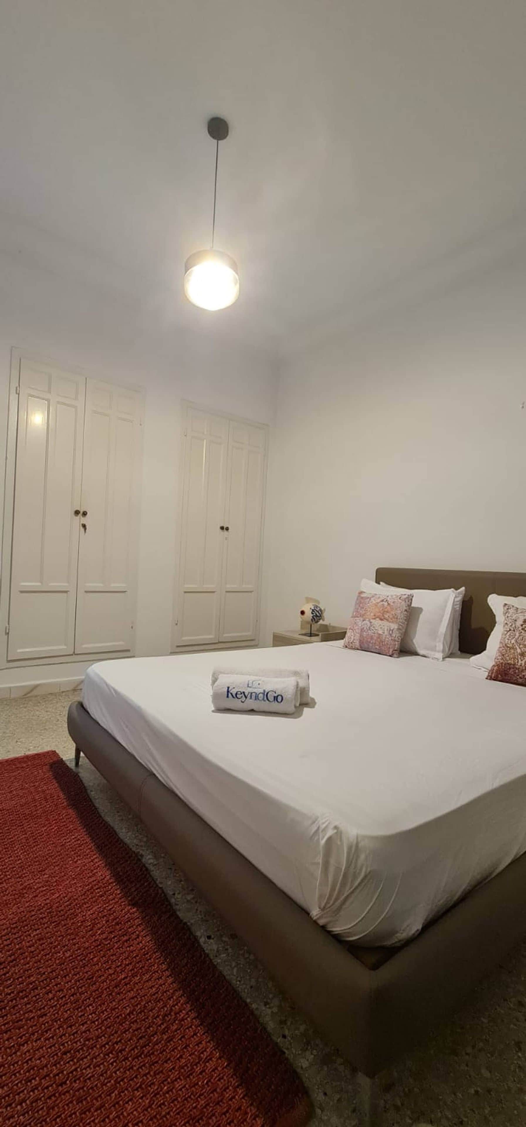 A bedroom with a bed, a red rug, white walls, a ceiling light, and a wardrobe.