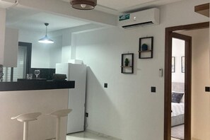 A modern kitchen with white cabinetry, a refrigerator, and a bar area with stools. There is a hallway leading to a bedroom with a bed and a nightstand. - Modern & Comfortable 1-Bedroom Apartment With Private Parking & Prime Location (Tunis)