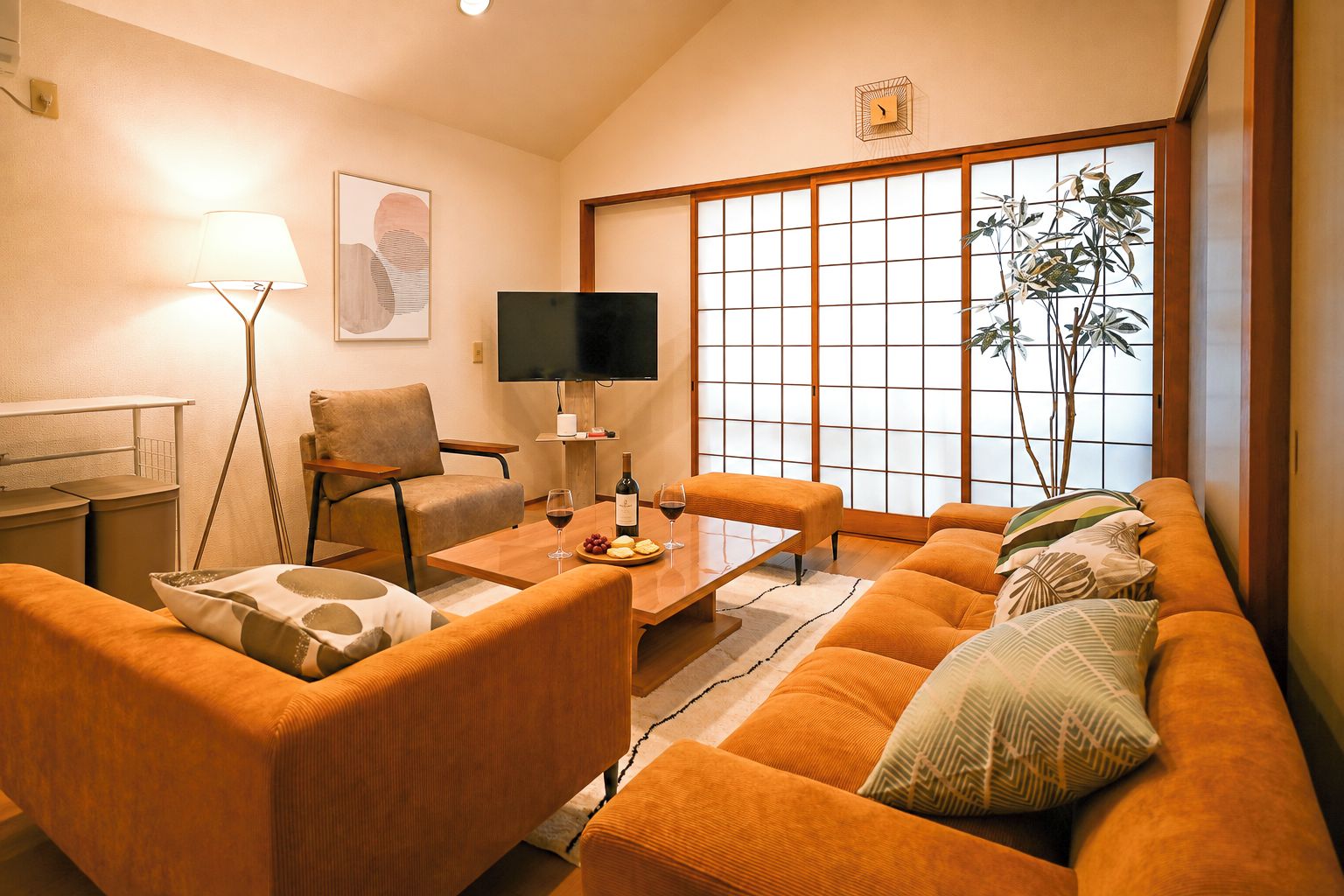 A living room with a sofa, armchairs, a coffee table, a TV, a lamp, and a large window with a view of a plant.