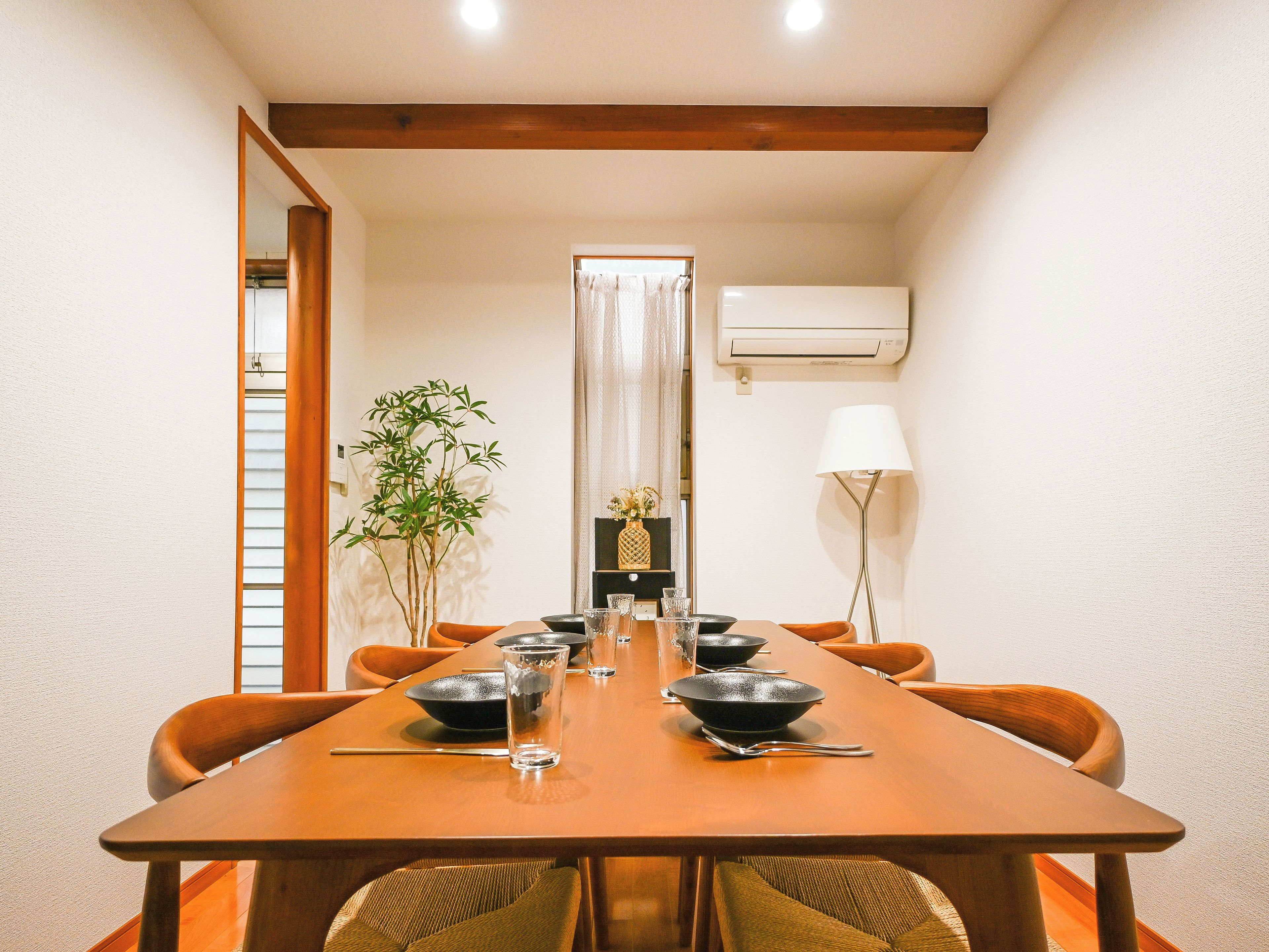 A dining area with a long table, chairs, a plant, and a lamp.