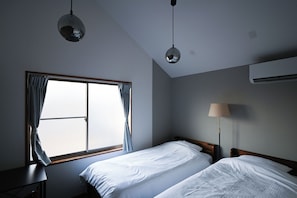 A hotel room with two beds, a window with curtains, a lamp, and an air conditioning unit. - Common Table Okayama - 5 beds & 5 bikes (岡山市)