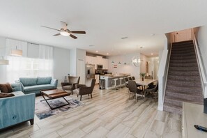 Ein modernes Wohnzimmer mit einer Couch, einem Couchtisch und Sesseln. Es gibt eine Küchenzeile mit einem Esstisch und Stühlen. Eine Treppe ist rechts sichtbar.