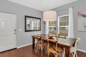 A dining area with a wooden table, chairs, a hanging light fixture, and a window with blinds. - Stylish 4BR Townhome 10 Min to Disney (Kissimmee)