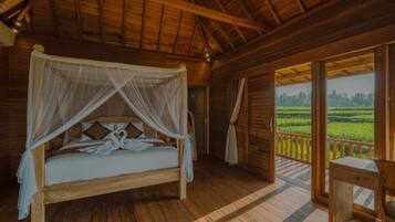 Una cama con dosel de madera y cuatro postes, un escritorio de madera, y vista a un paisaje verde a través de una puerta abierta.