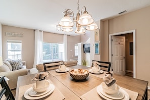 A dining area with a wooden table, white plates, and a bowl of food. There is a sofa and a TV in the background. - 3BR Bella Vida Pool Home by Walmart (Kissimmee)