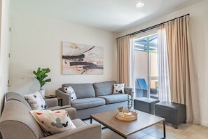 A modern living room with a grey sofa, a coffee table, and a large window with curtains. - 2BR Regal Oaks Townhome With Jacuzzi (Kissimmee)