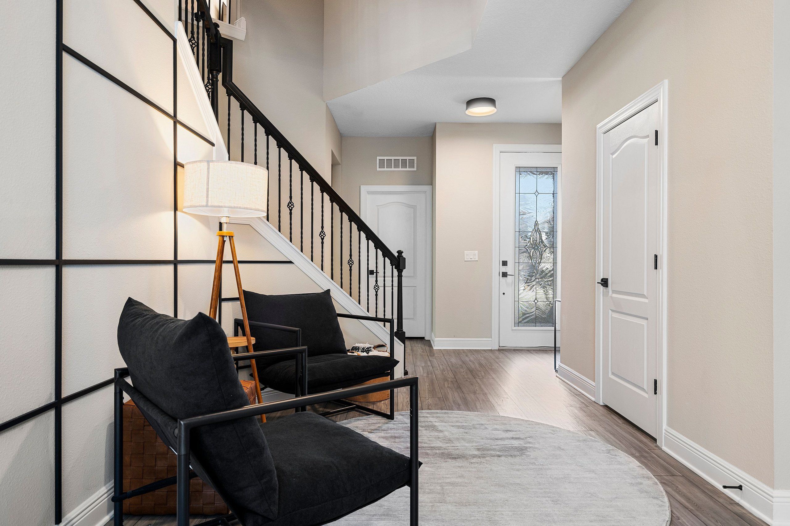 A hallway with a staircase, a lamp, two black chairs, and a rug.