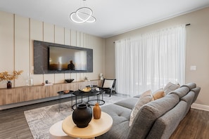 A modern living room with a large flat-screen TV mounted on the wall, a sectional sofa, a round coffee table, and a minimalist decor style. - Reunion 4BR Luxury Villa PS5 + Game Room (Kissimmee)