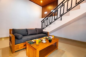 A wooden sofa with cushions, a wooden coffee table with food and drinks, and a staircase with a black railing. - Manikawana House by Dhananjaya (Ubud)