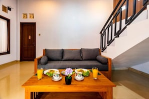 A room with a wooden table set for breakfast, a grey sofa, and a staircase. - Manikawana House by Dhananjaya (Ubud)