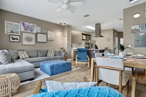 A modern living room with a grey sectional sofa, a blue ottoman, and a dining area with a wooden table and chairs. - Windsor Hills 3BR Pool,8min to Disney & Water Park (Kissimmee)