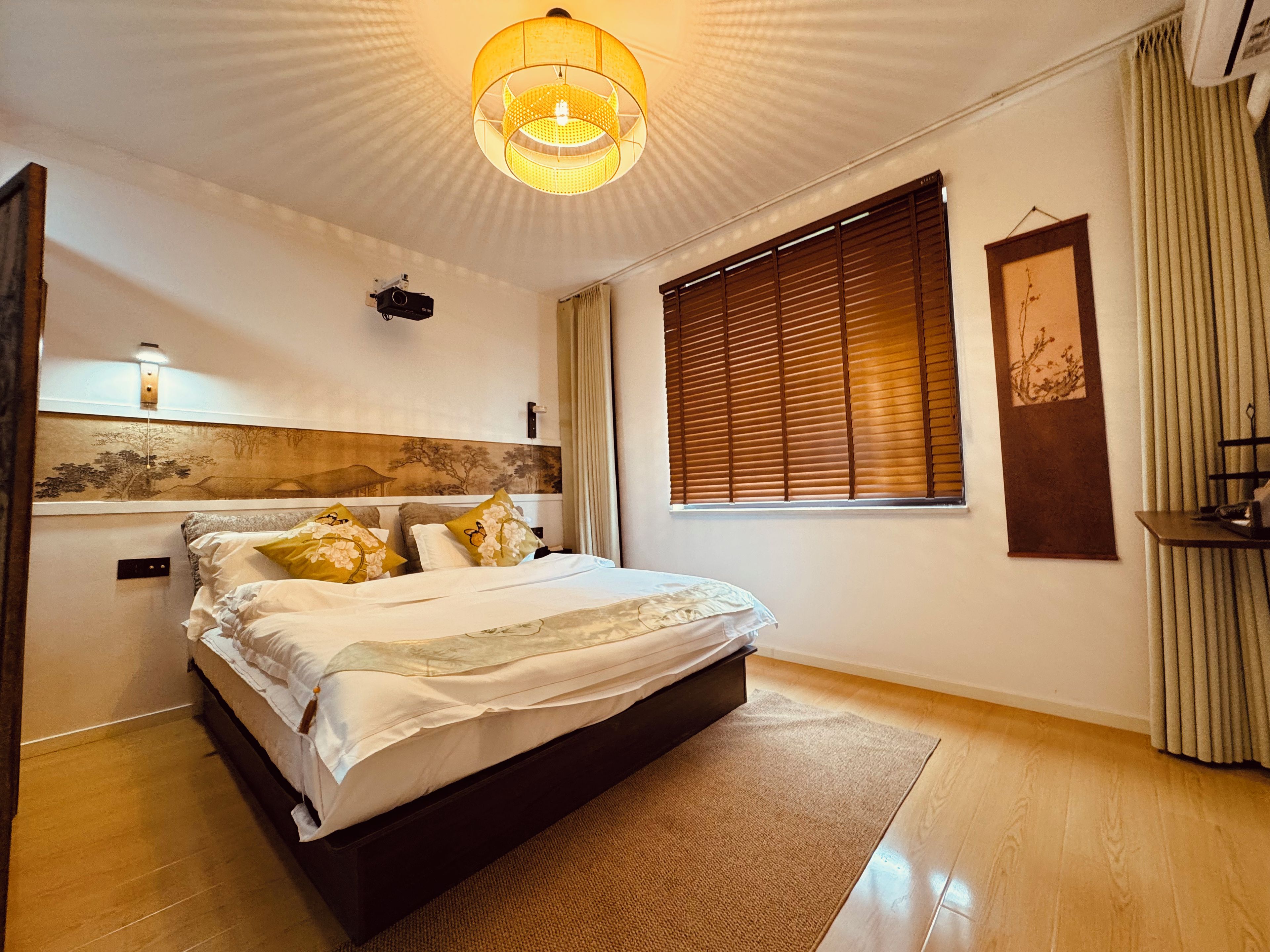 A bedroom with a bed, a wall-mounted projector, a large window with blinds, and a ceiling light fixture.