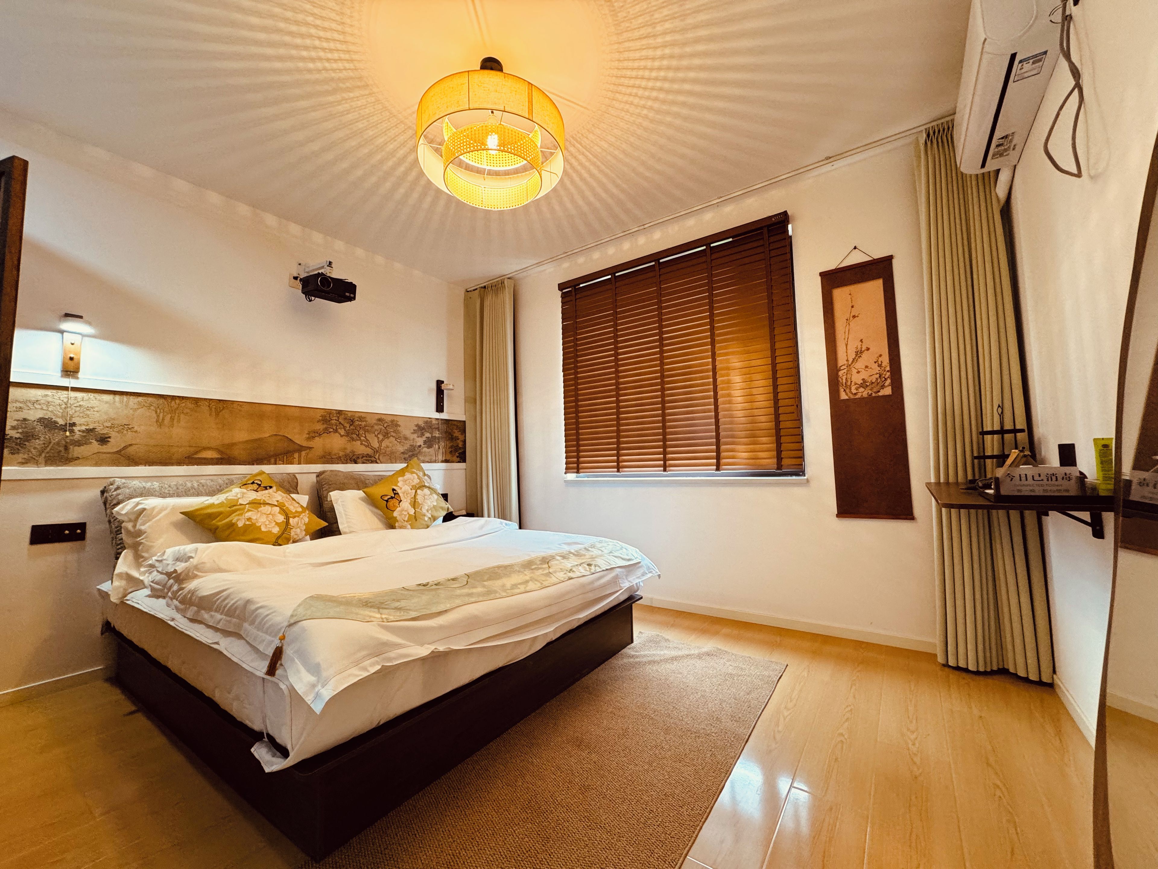 A modern bedroom with a large bed, a ceiling light, a window with blinds, and a bedside table.