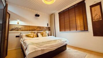 A bedroom with a bed, a wall-mounted light fixture, a window with blinds, and a framed artwork on the wall.