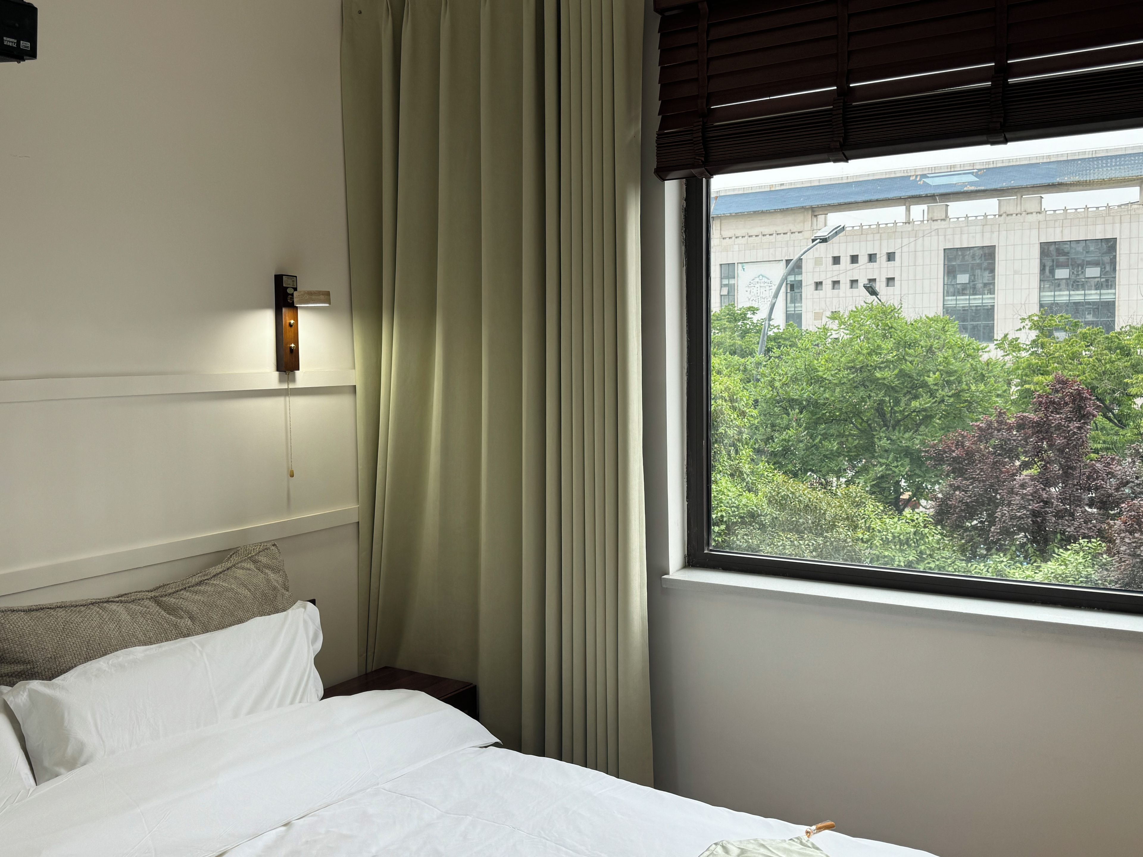Habitación de hotel con cama, ventana con persianas, luz de pared y vista a un edificio y árboles.