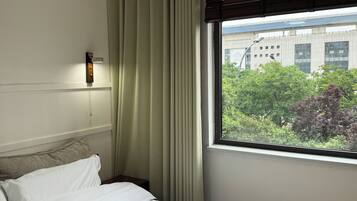 Habitación de hotel con cama, ventana con persianas, luz de pared y vista a un edificio y árboles.