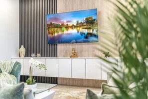 Ein modernes Wohnzimmer mit einem an einer Holzwand montierten Flachbildfernseher, einem weißen Schrank darunter und einem Glastisch mit einer Topfpflanze.