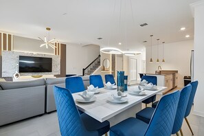 A modern dining area with a long table set for six, blue upholstered chairs, and a kitchen with a built-in TV and marble backsplash. - 5BR Storey Lake Townhome, Heated Pool (Kissimmee)