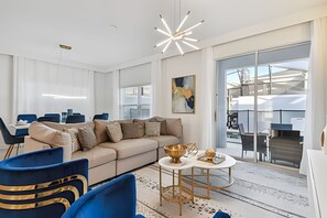 A modern living room with a sofa, blue chairs, a round table with a bowl, and a large window with a view of boats. - 5BR Storey Lake Townhome, Heated Pool (Kissimmee)