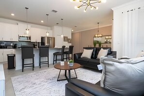 A modern living room with a sofa, chairs, a coffee table, and a kitchen area with white cabinets and appliances. - Villa Smile - 4bd/3ba Storey Lake Luxury Themed (Kissimmee)