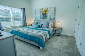 Ein Schlafzimmer mit einem Bett, zwei Nachttischen, einem Schreibtisch, einem Fenster mit Jalousien und einem großen abstrakten Gemälde an der Wand.