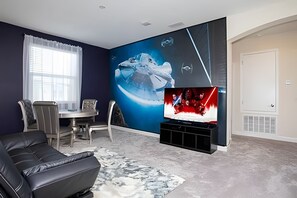 A modern living room with a large wall mural, a flat-screen TV, a leather sofa, and a dining area. - 5BR Storey Lake Home, Heated Pool (Kissimmee)