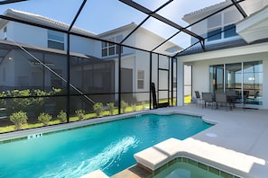 Une piscine intérieure moderne avec des parois en verre transparent, une terrasse ensoleillée dotée de mobilier d’extérieur et une vue sur les bâtiments voisins.